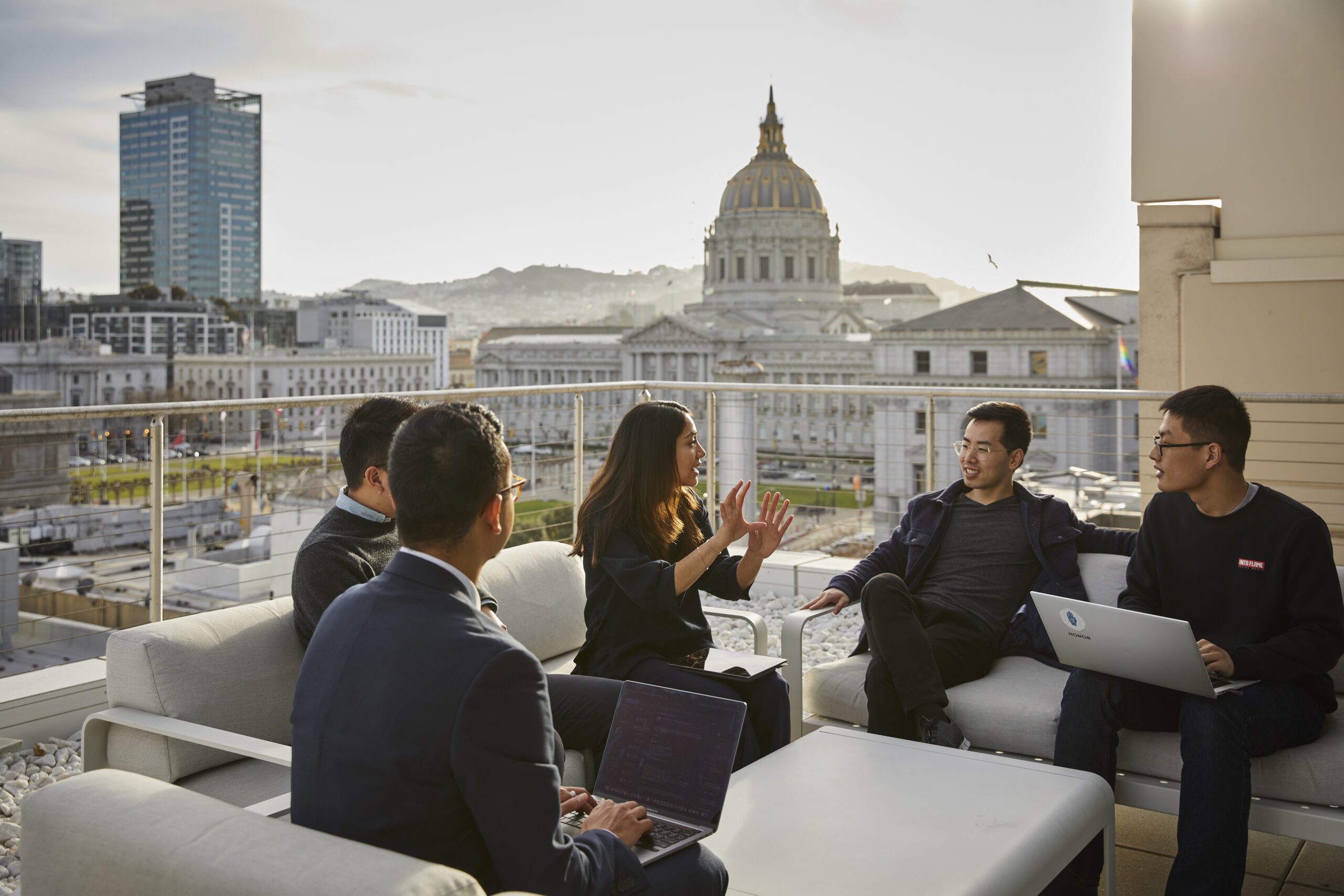 Group of individuals engaged in a discussion on a rooftop terrace overlooking San Francisco City Hall, with laptops on a coffee table.