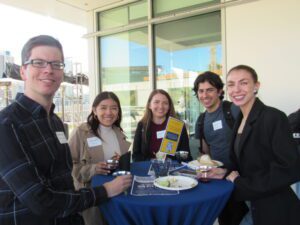 Law students engaging at a mixer event, standing around a table with food and drinks, smiling for the camera.