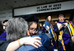 A graduate wearing glasses and graduation regalia hugs a loved one outside the commencement ceremony at Graham Auditorium.